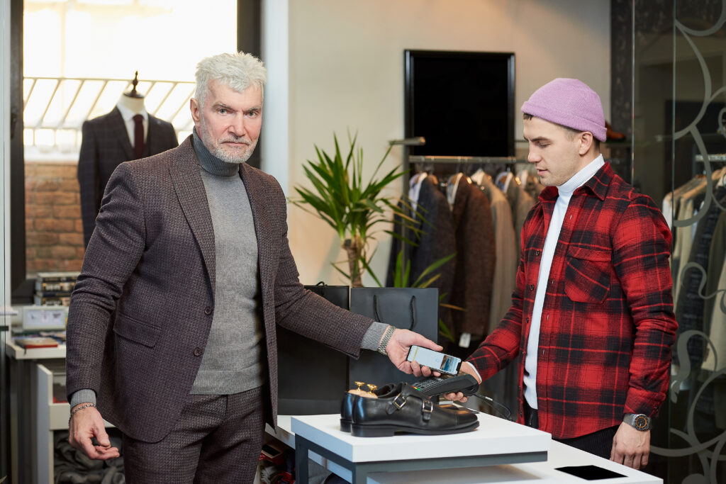 mature man with gray hair sporty physique is applying smartphone point sale terminal clothing store male customer with beard is paying shop assistant boutique